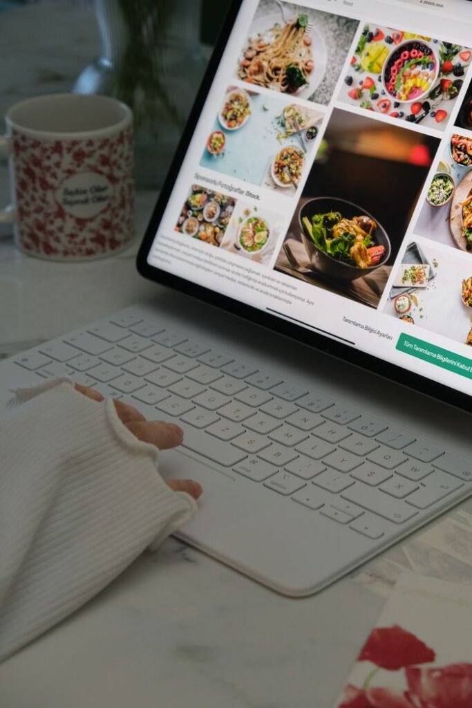 A person uses a white laptop to browse food recipes, with a mug and cozy atmosphere.